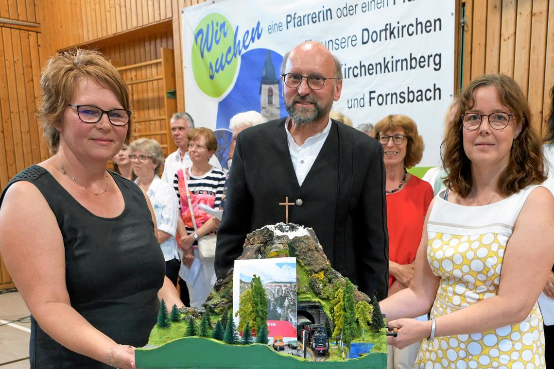 Die Kirchengemeinderätinnen Cornelia Lange (links) und Antje Noller (rechts) halten für Steffen Kaltenbach ein liebevoll gestaltetes Abschiedsgeschenk bereit: ein Modelleisenbahn-Bergdiorama mit Gutschein für eine Bergbahnfahrt in der Schweiz. Foto: Elisabeth Klaper