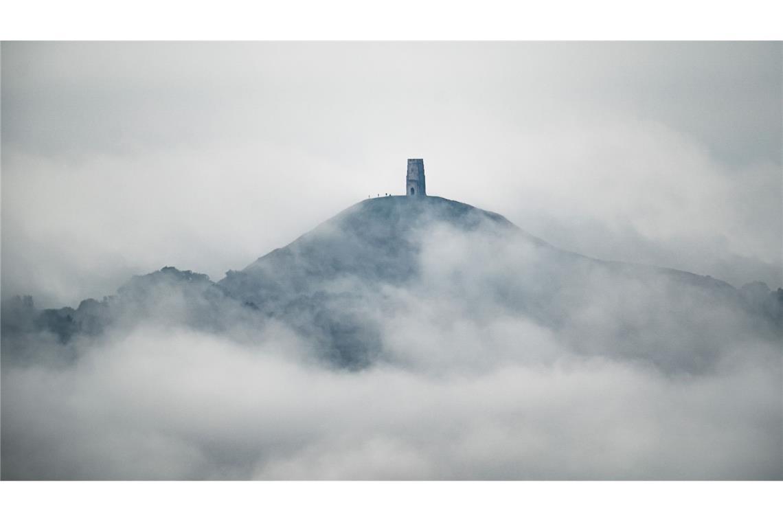 Eine Kulisse wie aus einer Fantasyserie: Der St. Michael's Tower im englischen Glastonbury ist in Morgennebel und Wolken gehüllt. Am alten Tor auf dem Hügel stehen Wanderer.