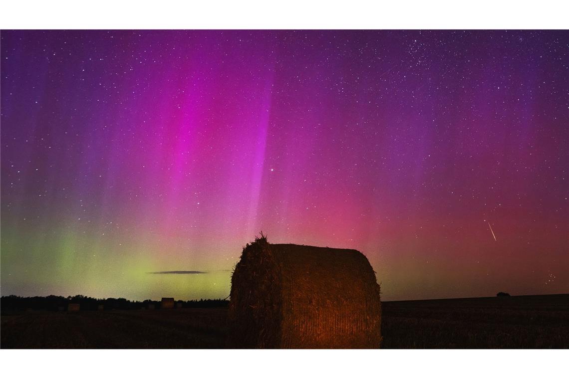 Nordlichter am Himmel, während eine Sternschnuppe über  einem Strohballen am Himmel verglüht.