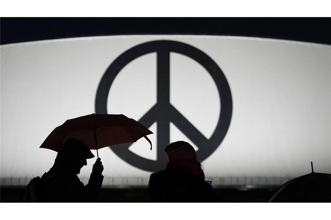 Passanten gehen mit Regenschirmen vorbei an einem leuchtenden Peace-Zeichen im strömenden Regen zum Musical-Theater "Stage Theater an der Elbe" im Hafen.